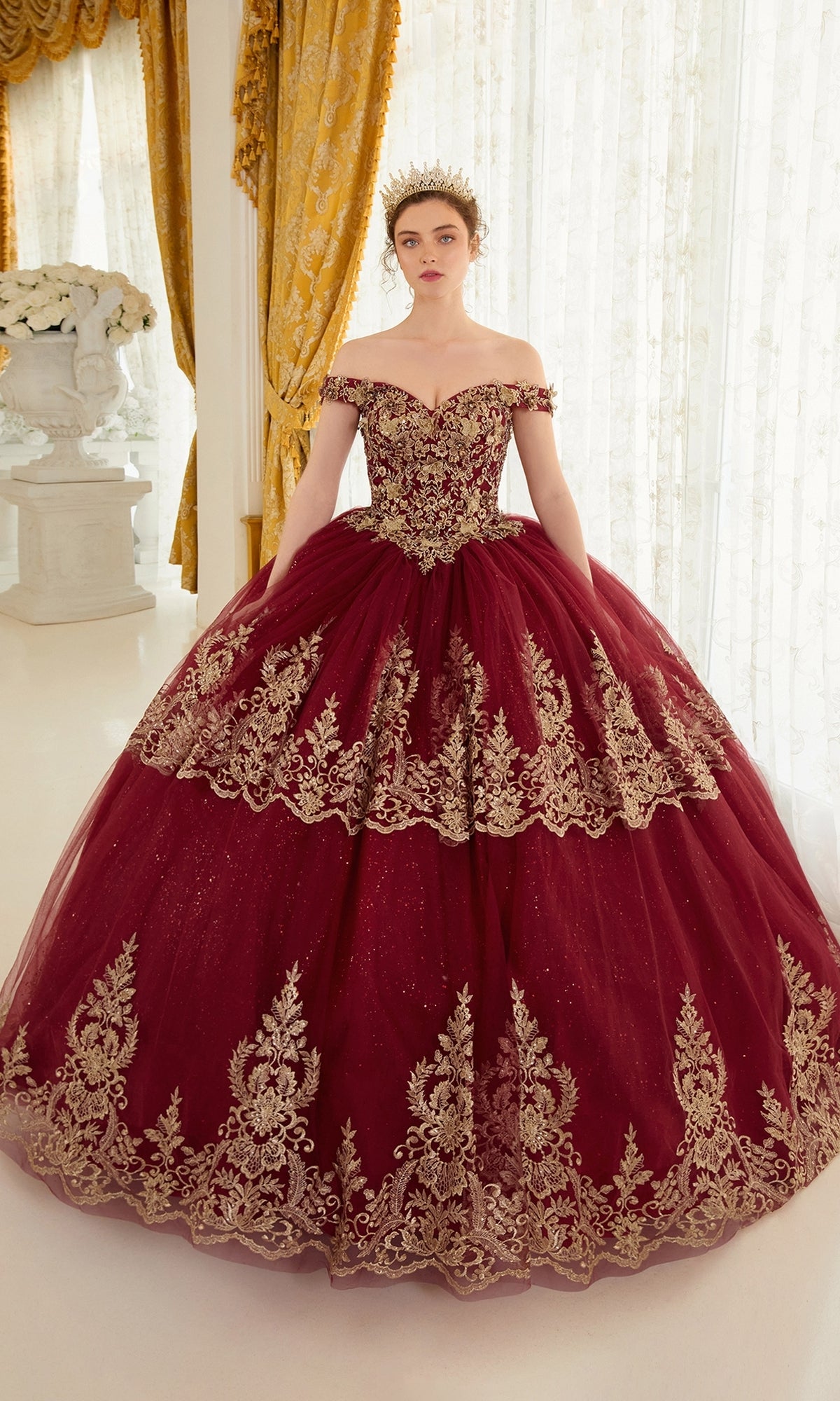 A woman stands in an elegant room with gold curtains and white decor, wearing the Ladivine 15705 Quince Dress featuring gold embroidery and an off-the-shoulder design, topped with a gold crown.